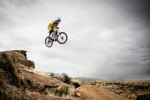 A cyclist performs a daring jump off a rocky cliff under a cloudy sky.